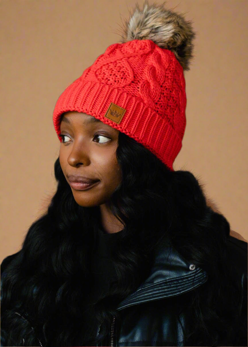 Person wearing a red knitted beanie with a fur pom-pom on a black background