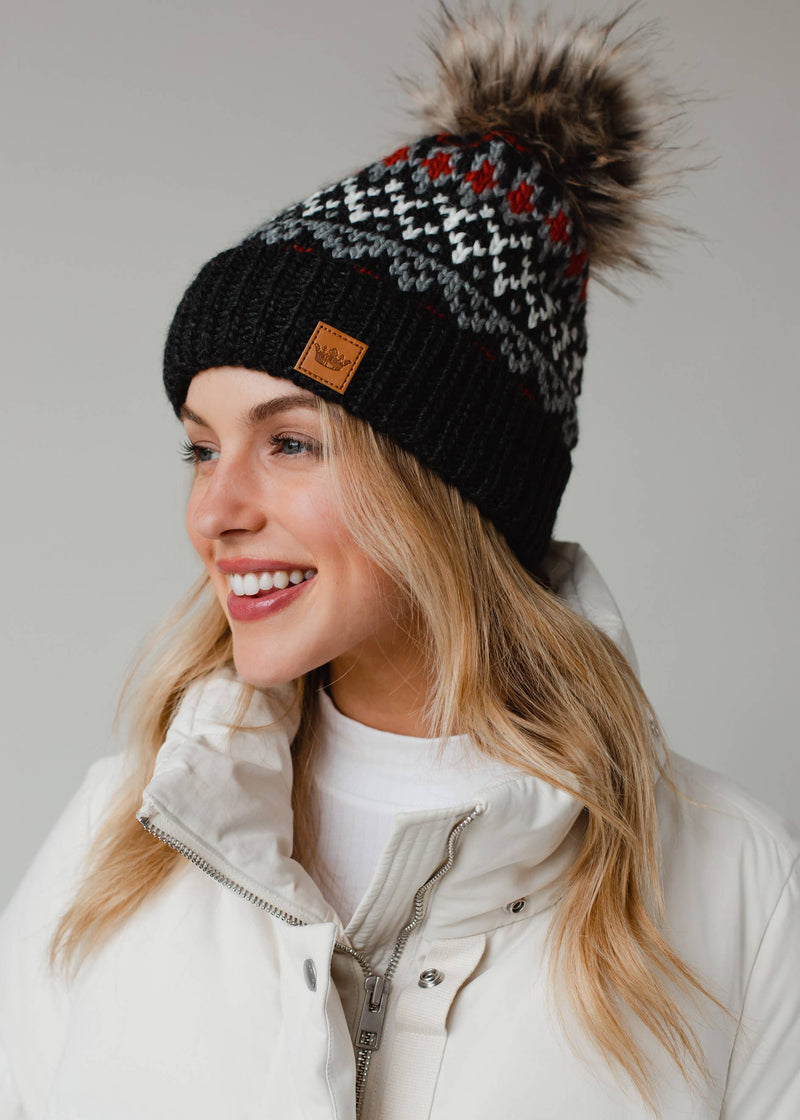 Woman wearing a black knit beanie with a pom-pom and a visible brand logo, smiling against a neutral background.
