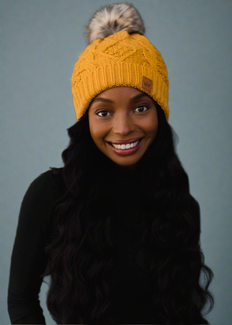 Woman wearing a yellow knit beanie with a fur pom-pom against a black background
