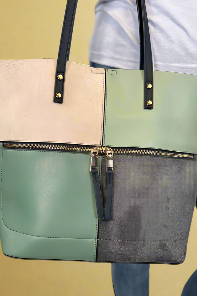 Model is carrying a large color block tote bag with external zippers in olive sage green. 