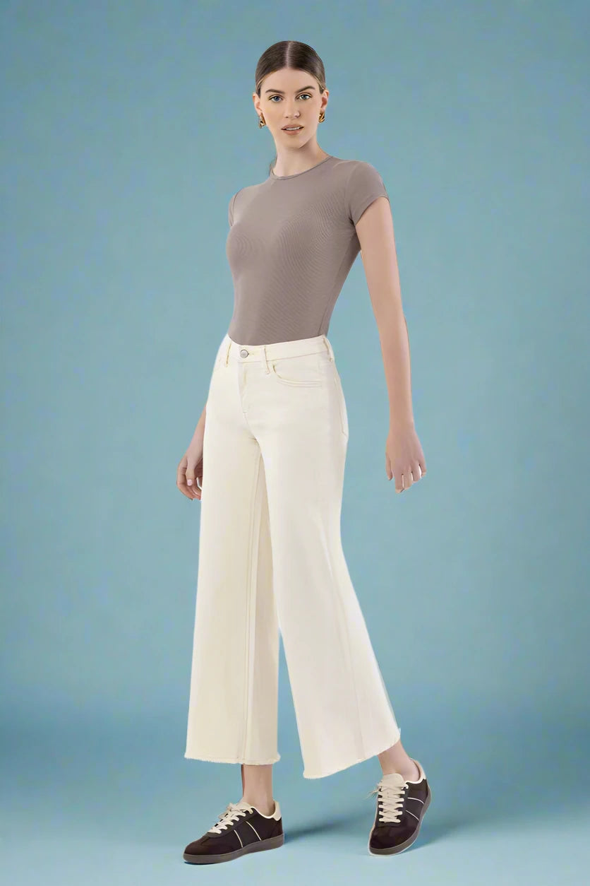 Woman wearing a brown top and white wide leg pants on a plain background.