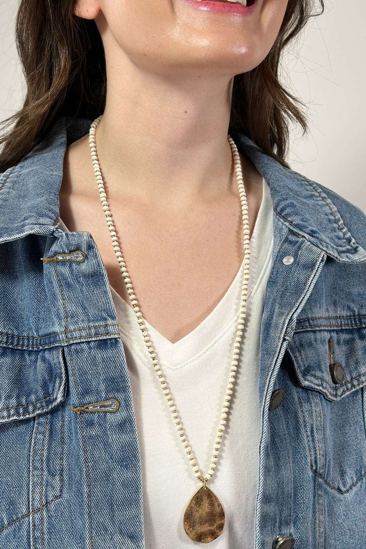 Model is wearing a long beaded necklace with gold teardrop pendant. 