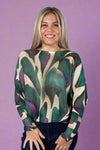Model is wearing a light knit sweater with dolman sleeves and a vibrant dark teal and purple pattern. 