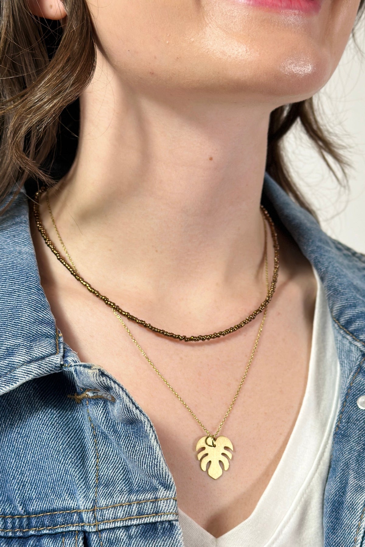 Model is wearing a two layer necklace with brown beads and gold leaf pendant. 