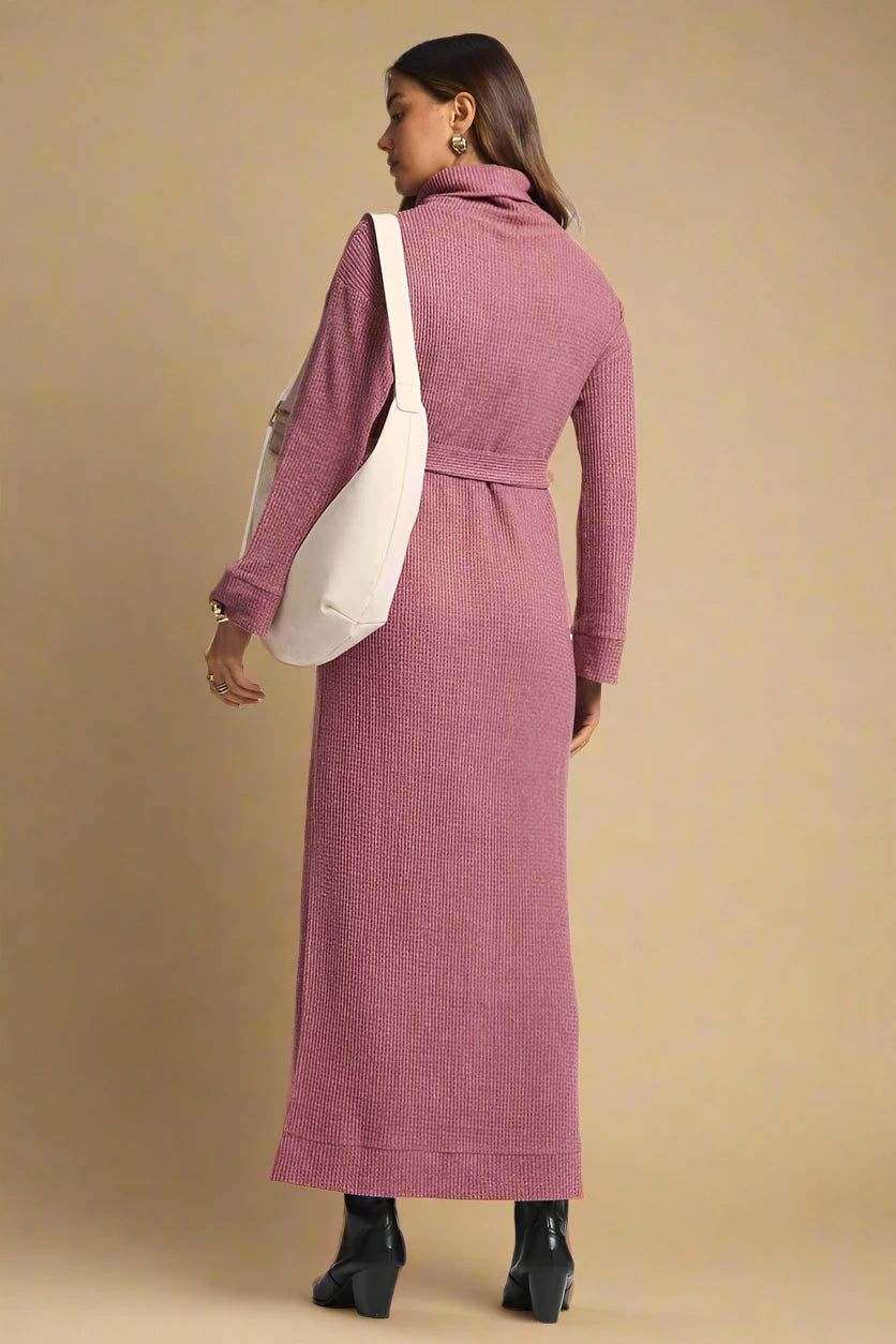 Woman wearing a long pink dress with a white bag on a plain background.