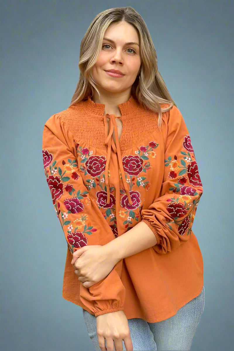A marigold orange, smocked tie neckline blouse with floral embroidery.
