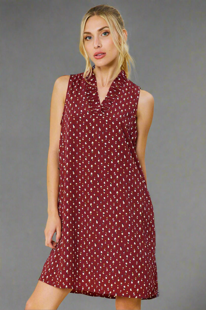 Model is wearing a burgundy sleeveless dress with an abstract print. The dress features a v neck with a ruffle detail. 