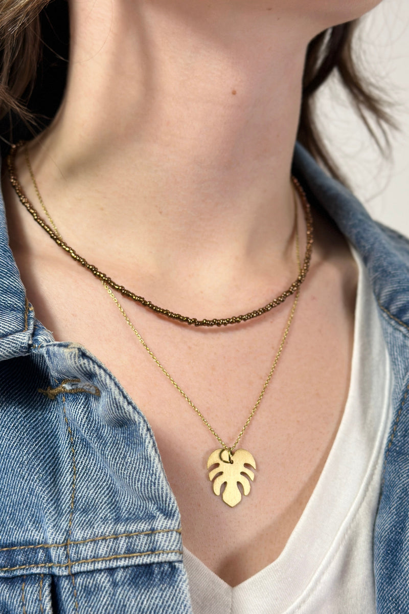 Model is wearing a two layer necklace with brown beads and gold leaf pendant. 