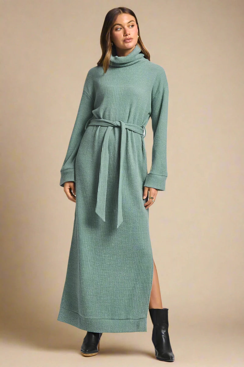 Woman wearing a long green dress with a belt on a plain background