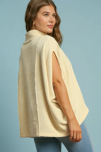 Woman wearing a two-tone cape over a cream top and blue jeans on a brown background