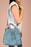 Model is holding a blue quilted bucket bag with handle and cross body strap. Purse includes removable wristlet.