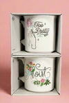 White ceramic mugs with funny phrases. One cup says “Too Pretty for Prison” and the other says “Moist”.