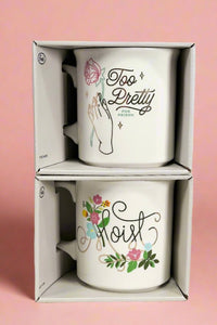 White ceramic mugs with funny phrases. One cup says “Too Pretty for Prison” and the other says “Moist”.