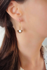 A white agate stone pendant surrounded by gold sunbursts, attached to a thin gold hoops.