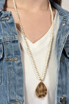 Model is wearing a long beaded necklace with gold teardrop pendant. 