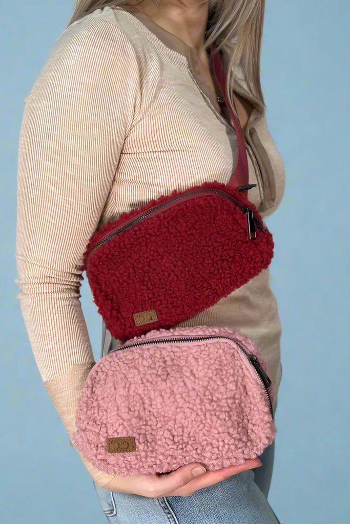 Model showcases the Sherpa Belt Fanny Pack in both burgundy and rose. Each bag features a vinyl chest/waist strap, zipper closure, and soft, Sherpa body.