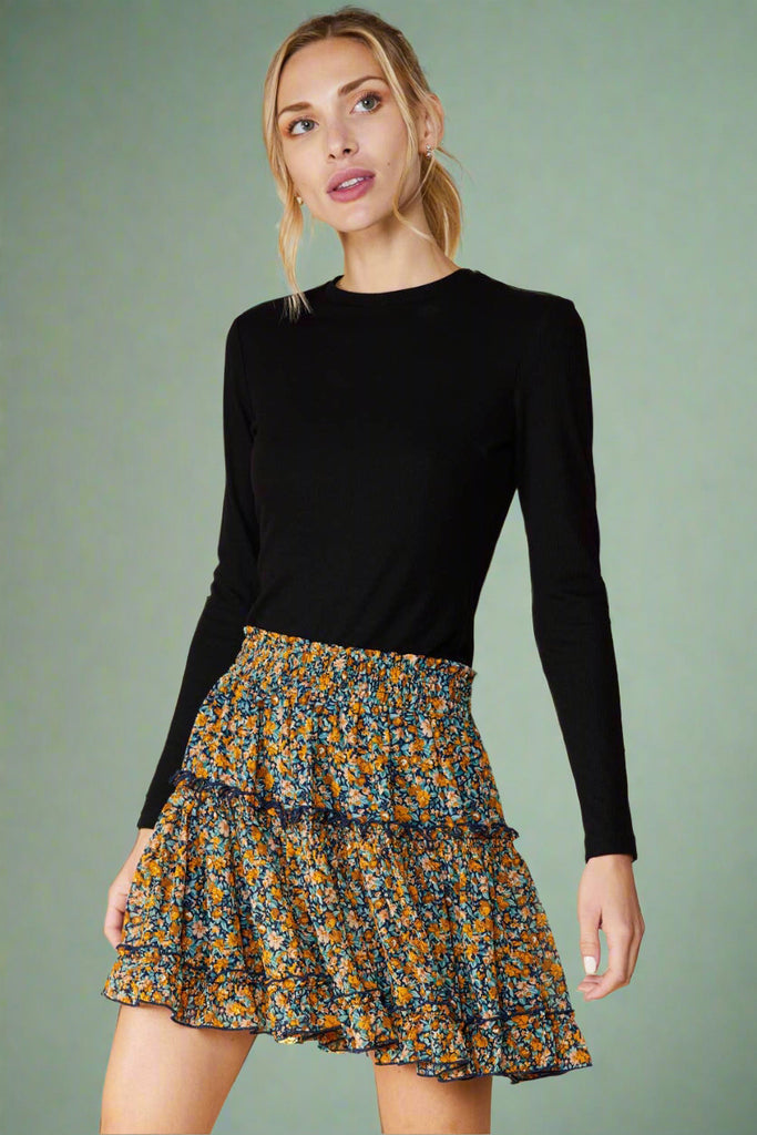 Model is wearing a floral mini skirt featuring an elastic waistband. The skirt is navy blue with an all over floral print in mustard yellow. It is fully lined and has ruffled details. 