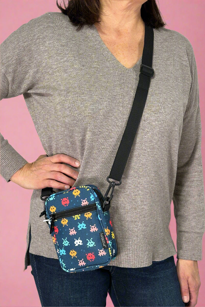 Model is wearing a small crossbody bag with a Pac-Man inspired print with an external zipper pocket and adjustable canvas strap. 