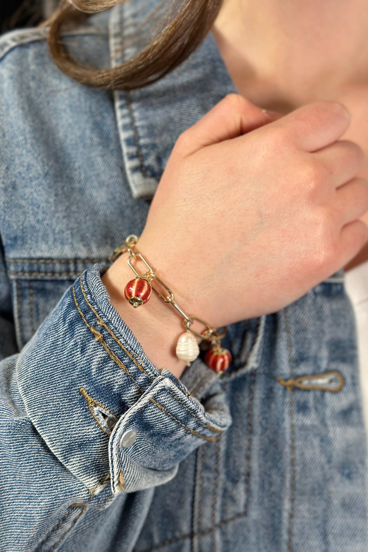 Model is wearing a gold paperclip chain bracelet with red and white charms. 