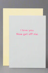 A greeting card with solid off-white front and bright pink text that reads ‘I love you. Now get off me.’