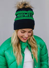 Person wearing a black and green 'Midwest' beanie with a fur pom-pom, standing against a gray background.