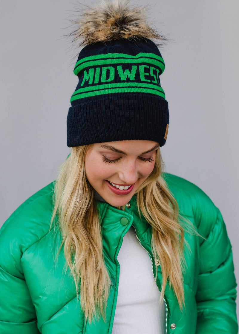 Person wearing a black and green 'Midwest' beanie with a fur pom-pom, standing against a gray background.