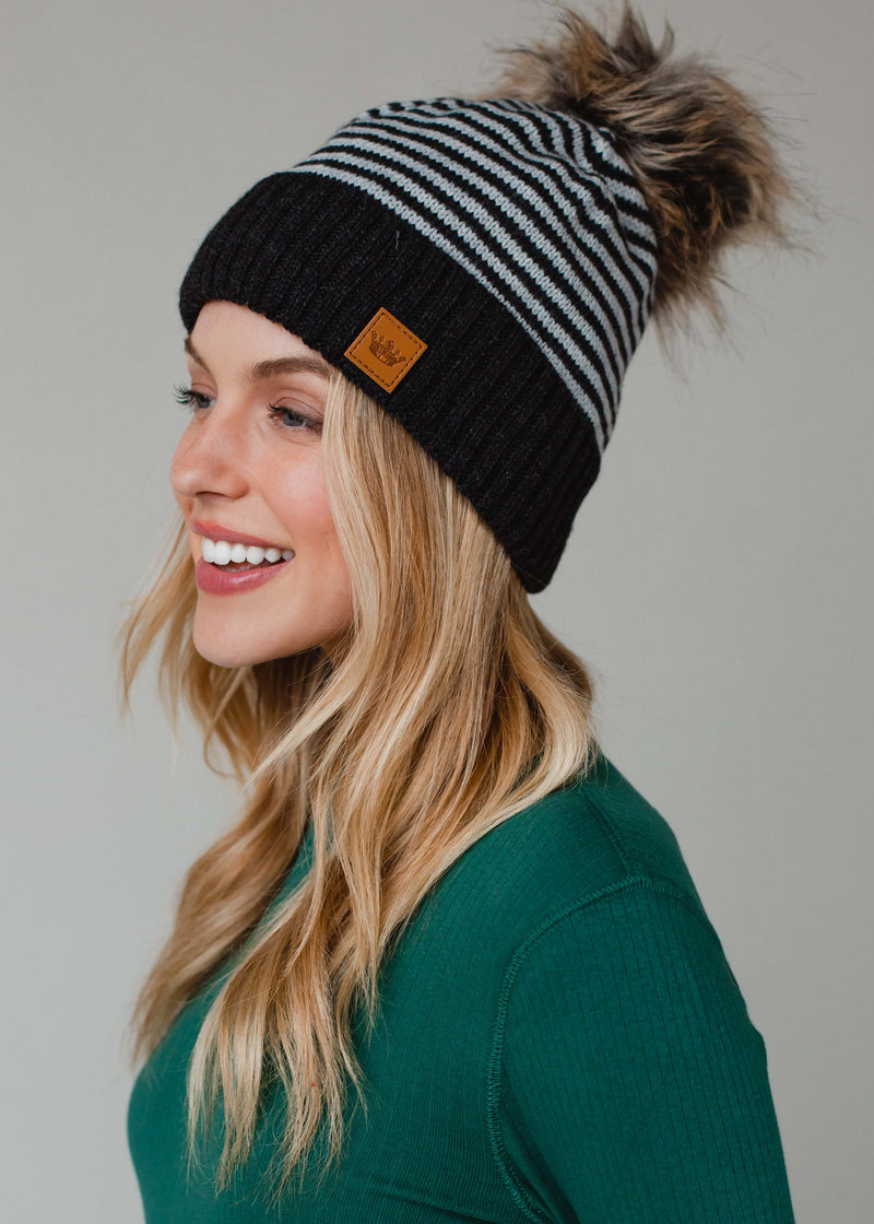 Woman wearing a black and gray striped beanie with a pom-pom on a plain background