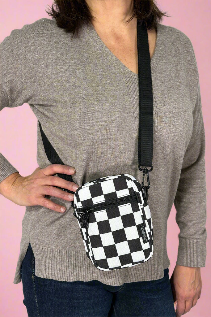 Model is wearing a small black and white checkered crossbody bag with an external zipper pocket and adjustable canvas strap. 