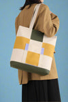 Models holds adorable, canvas shoulder bags featuring a modern, blocky, checkered plaid with green, yellow, and lavender accents.