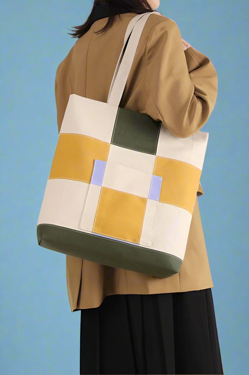 Models holds adorable, canvas shoulder bags featuring a modern, blocky, checkered plaid with green, yellow, and lavender accents.