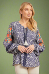 A blue and white floral pattern blouse with a tie neckline. The sleeves boast colorful embroidered flowers. Navy lace accentuates this top.