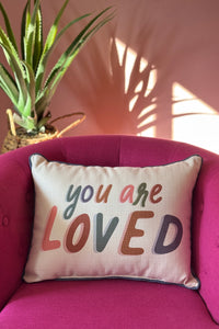 A beige rectangle pillow that has blue piping edge and says ‘You Are Loved’ on the front.
