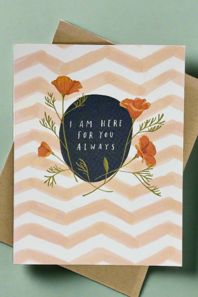 A greeting card with white and peach colored chevron style stripes with black circle surrounded by flowers and text that reads ‘I am here for you always.’