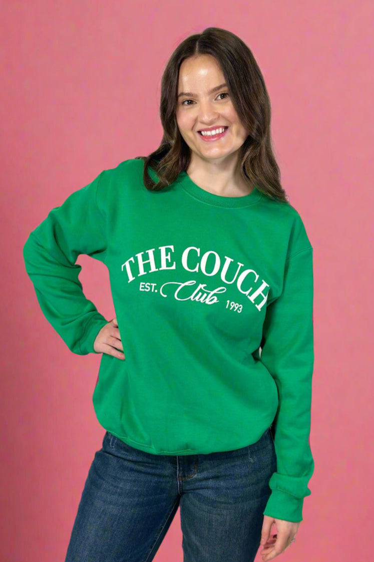 Model is wearing a kelly green colored fleece lined crew neck with the phrase "The Couch Club" printed across the front in velvet lettering. 