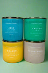 Four candles displayed, each with a different scent and color glass container. The container colors and scents include blue glass in Aqua Juniper, green glass in Cactus Rain, yellow glass in golden amber and tan glass in sandalwood bonfire.