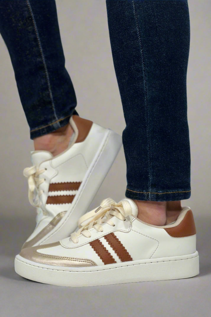 Model is wearing tan and white soccer style sneakers with two tan stripes and pop of gold. 