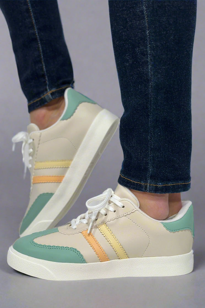 Model is wearing off-white and mint colored soccer style sneakers with an orange and yellow stripe on the side panel. 