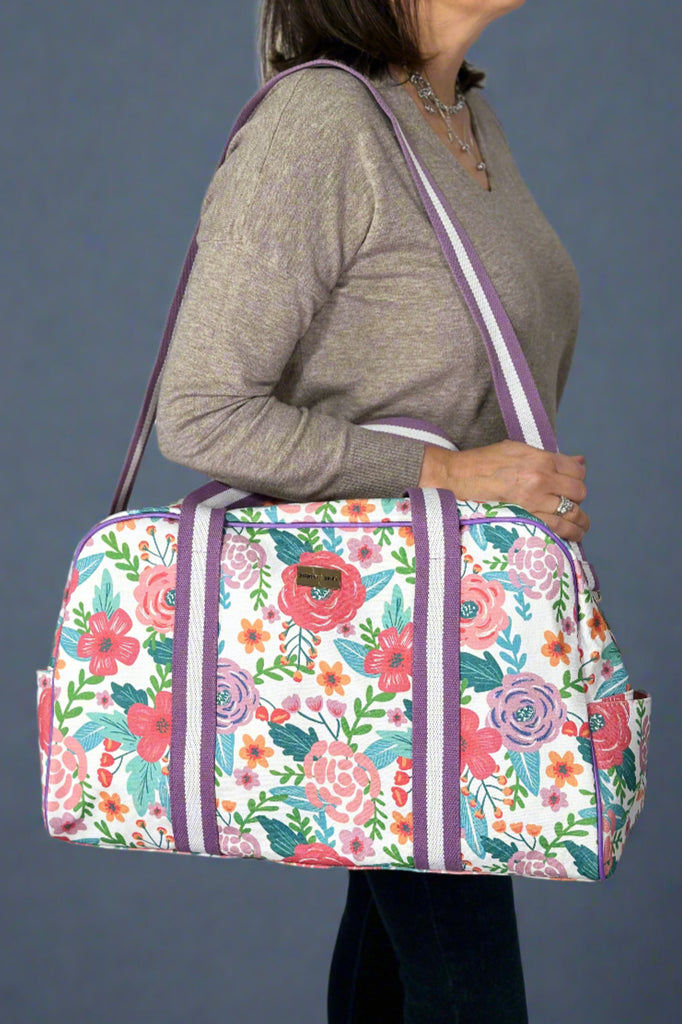 Model is holding a floral printed canvas duffle bag with top handles and a removable shoulder strap. 