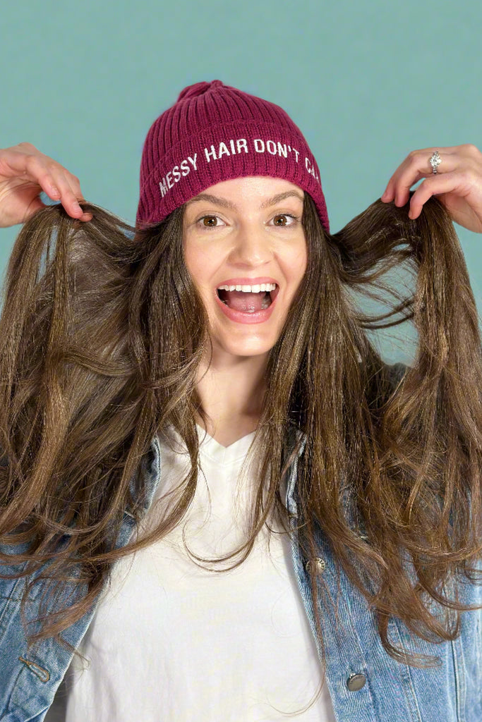 Model is wearing a berry colored ribbed knit beanie that has the phrase "MESSY HAIR DON'T CARE" embroidered across the front in white.