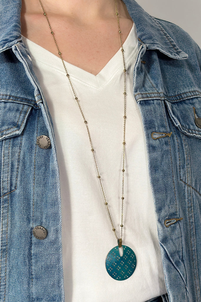 model is wearing a long necklace with a teal colored pendant. 