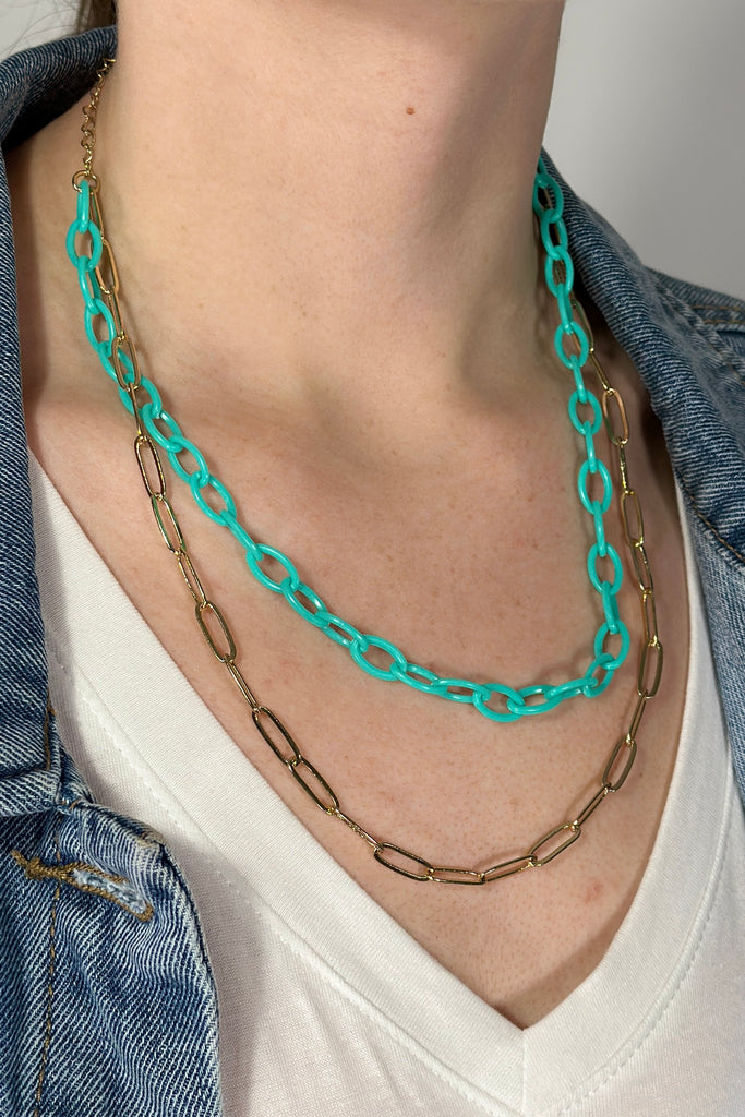 Model is wearing a double chain necklace. One is gold chain links and the other is teal acrylic chain links.