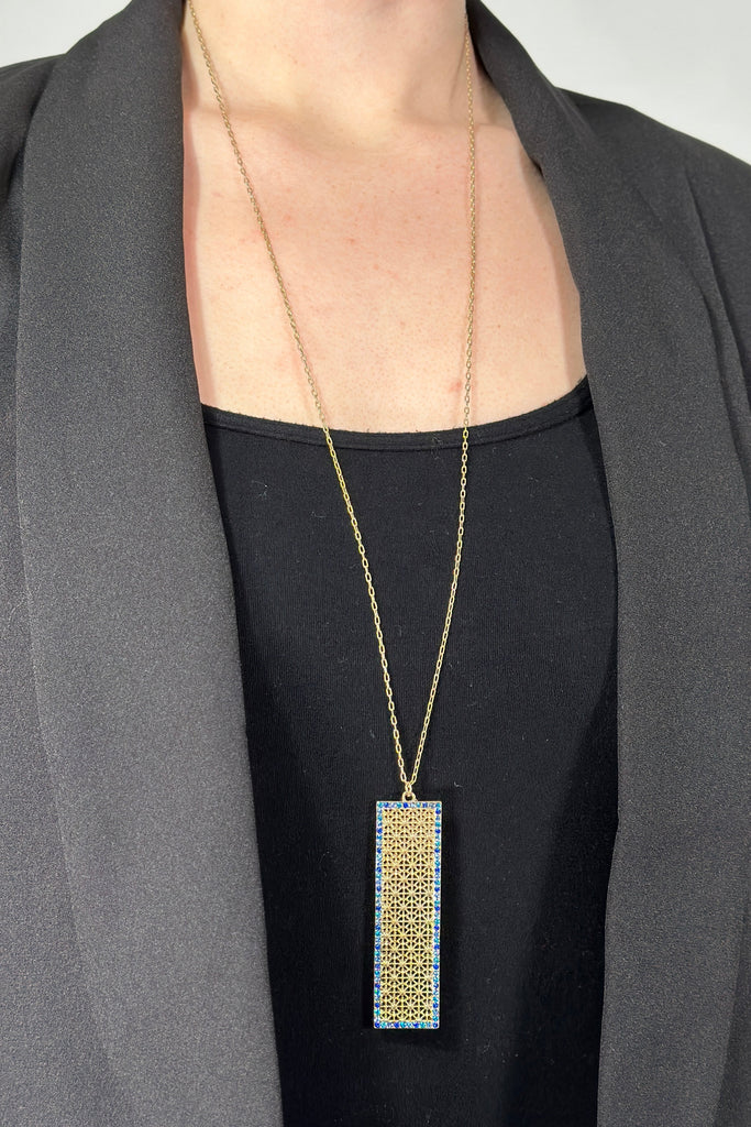 Model is wearing a long gold necklace with a rectangular pendant. 