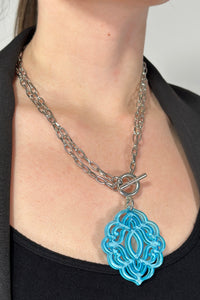 Model is wearing a silver chain necklace that features a blue acrylic pendant. 