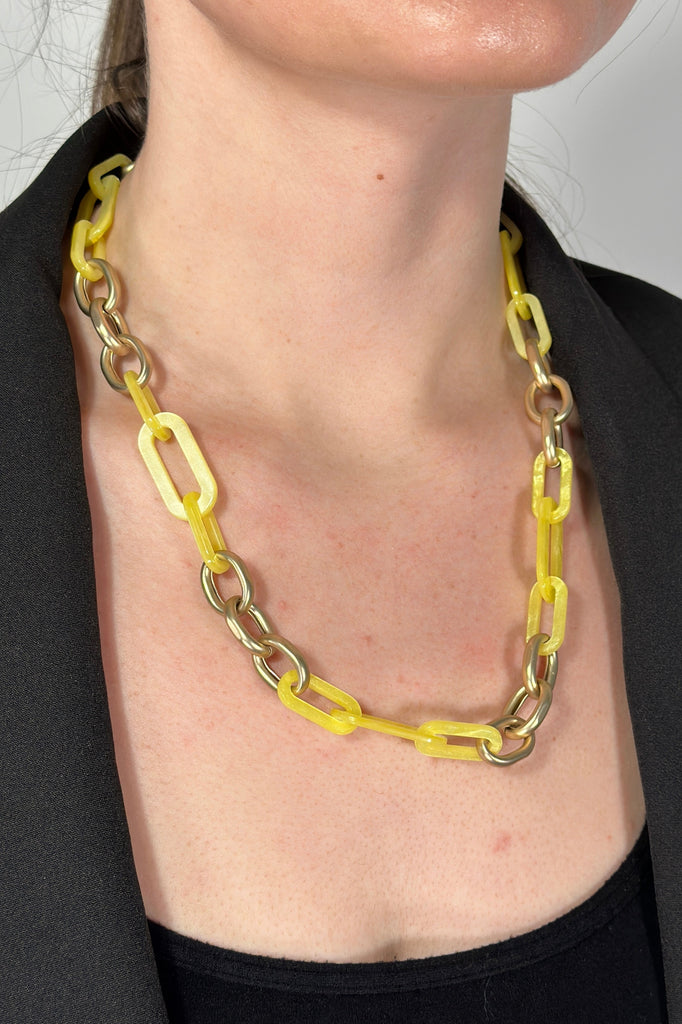 Model is wearing a chunky acrylic chainlink necklace that is gold with neon yellow links. 