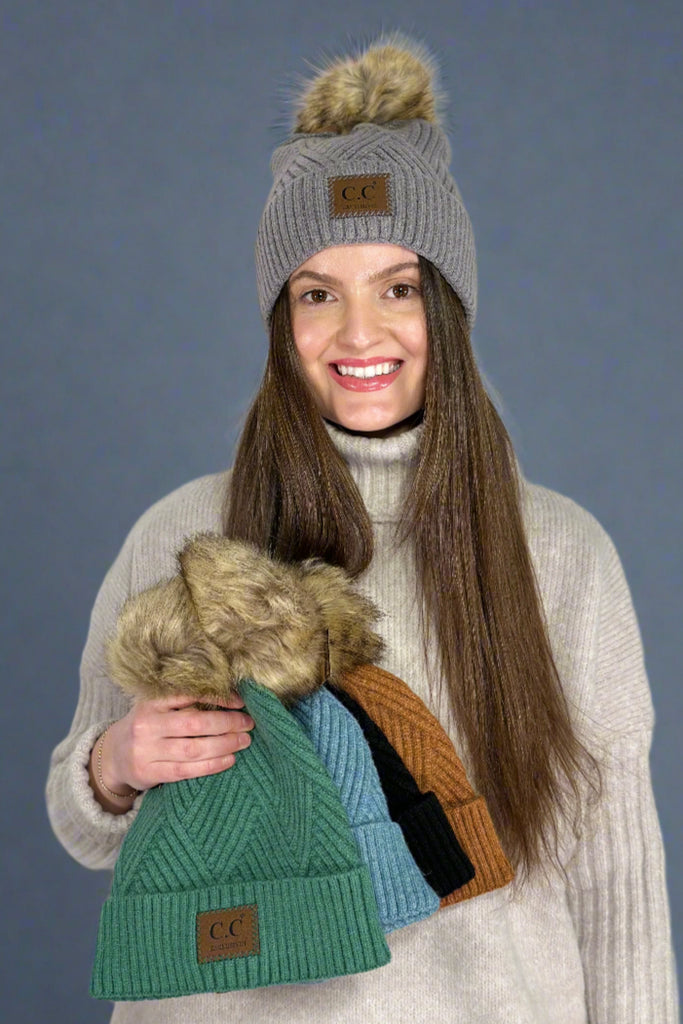 Model is wearing geometric patterned beanie with a fur pom and holding the same hat in four other colors. 