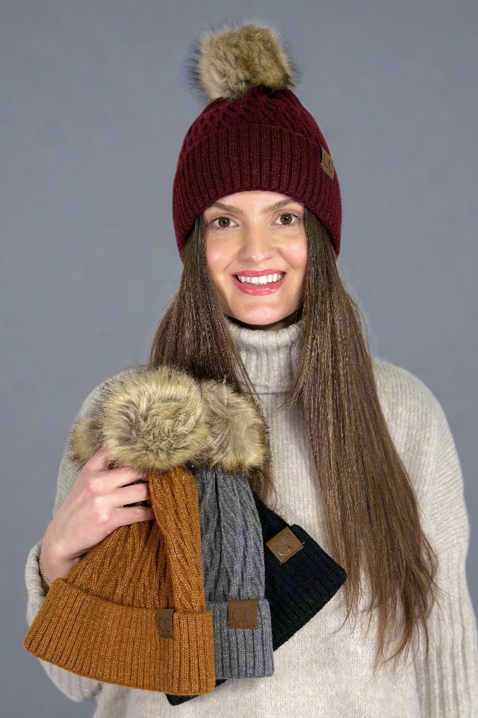 Model is wearing a honey comb patterned beanie with a fur pom. She's holding three others available in different colors. 