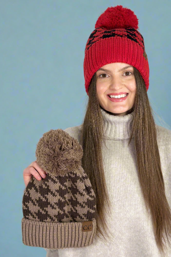 Model is wearing a houndstooth printed beanie with a pom and holding the other color available in her hand.