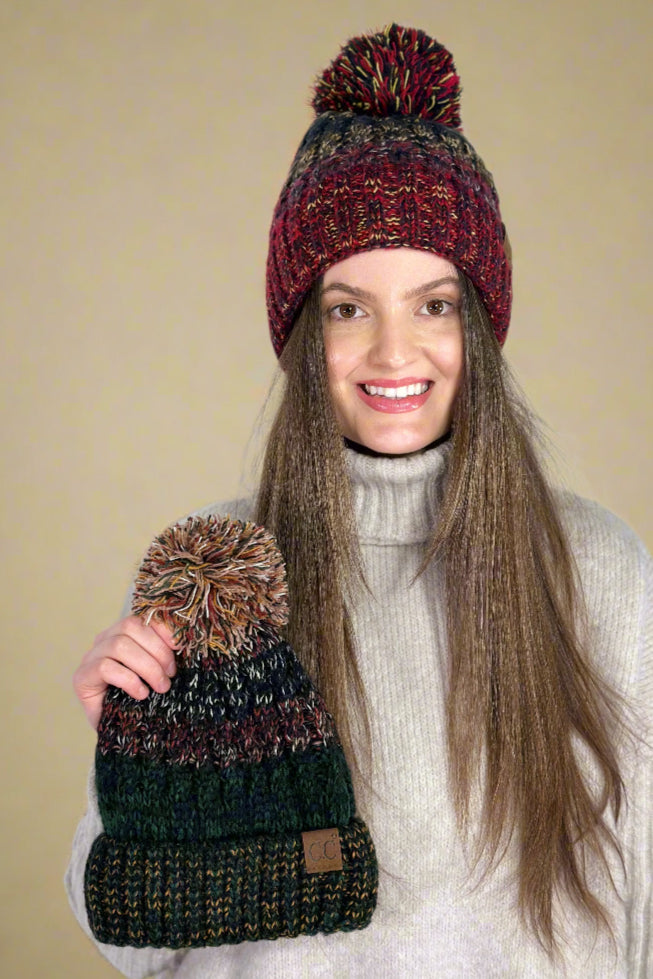 Model is wearing a cuffed multicolor pom beanie and holding another color option of the hat in her hand.