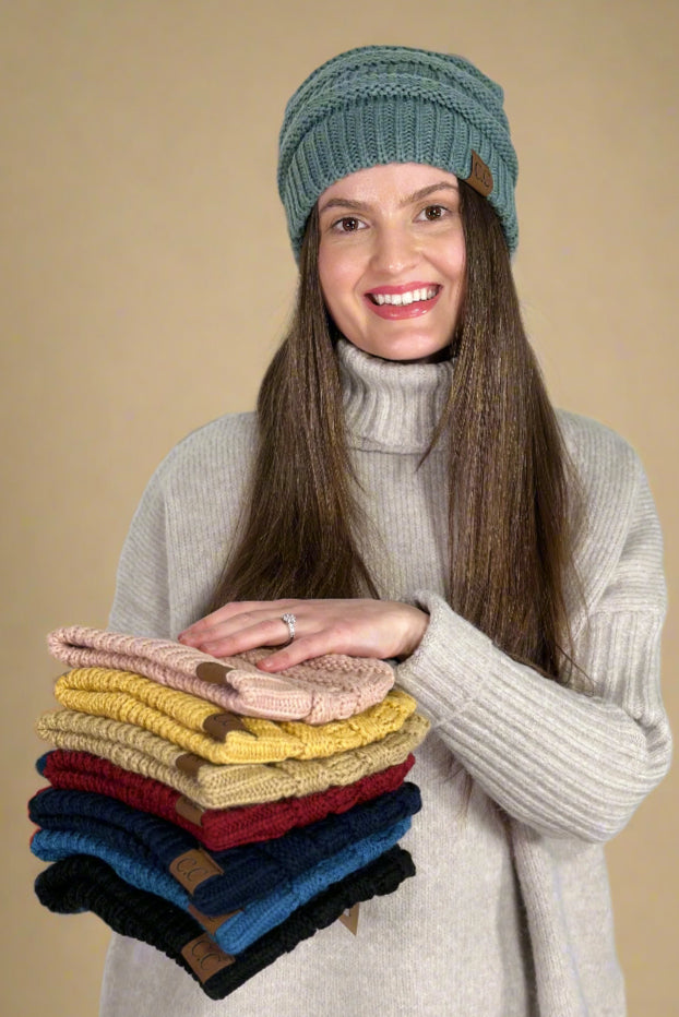 Model is wearing a classic cable knit beanie and holding a stack of beanies showcasing all the available colors. 