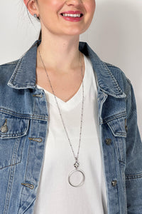 Model is wearing a long silver chain necklace with a small clover pendant that has a large hoop attached to it. 
